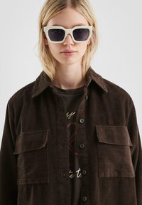 Braune Cordjacke mit zwei Brusttaschen, kombiniert mit einem dunkelbedruckten T-Shirt. Weiße quadratische Sonnenbrille auf dem Kopf.