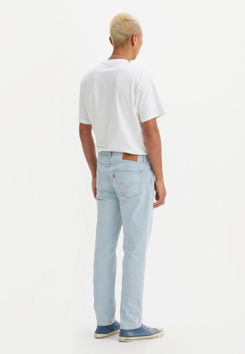 Jean en denim bleu clair avec une coupe droite, patch en cuir à l'arrière de la taille et coutures discrètes. Porté avec un t-shirt blanc et des baskets bleues.