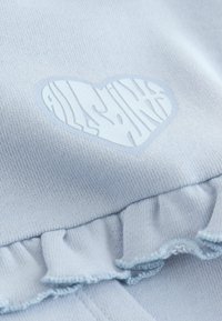 Material tricotat albastru deschis cu un logo în formă de inimă și text stilizat. Un tiv cu volane adaugă textură și detalii designului.