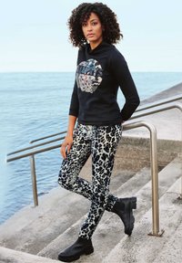 Schwarzer Hoodie mit grafischem Druck, kombiniert mit grauen und schwarzen Leopardenmuster-Hosen. Model trägt schwarze Ankle Boots und steht auf Betonstufen.