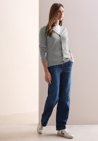 Cardigan gris clair avec des boutons, associé à un jean bleu foncé. Le modèle porte des baskets blanches, sur un fond neutre.