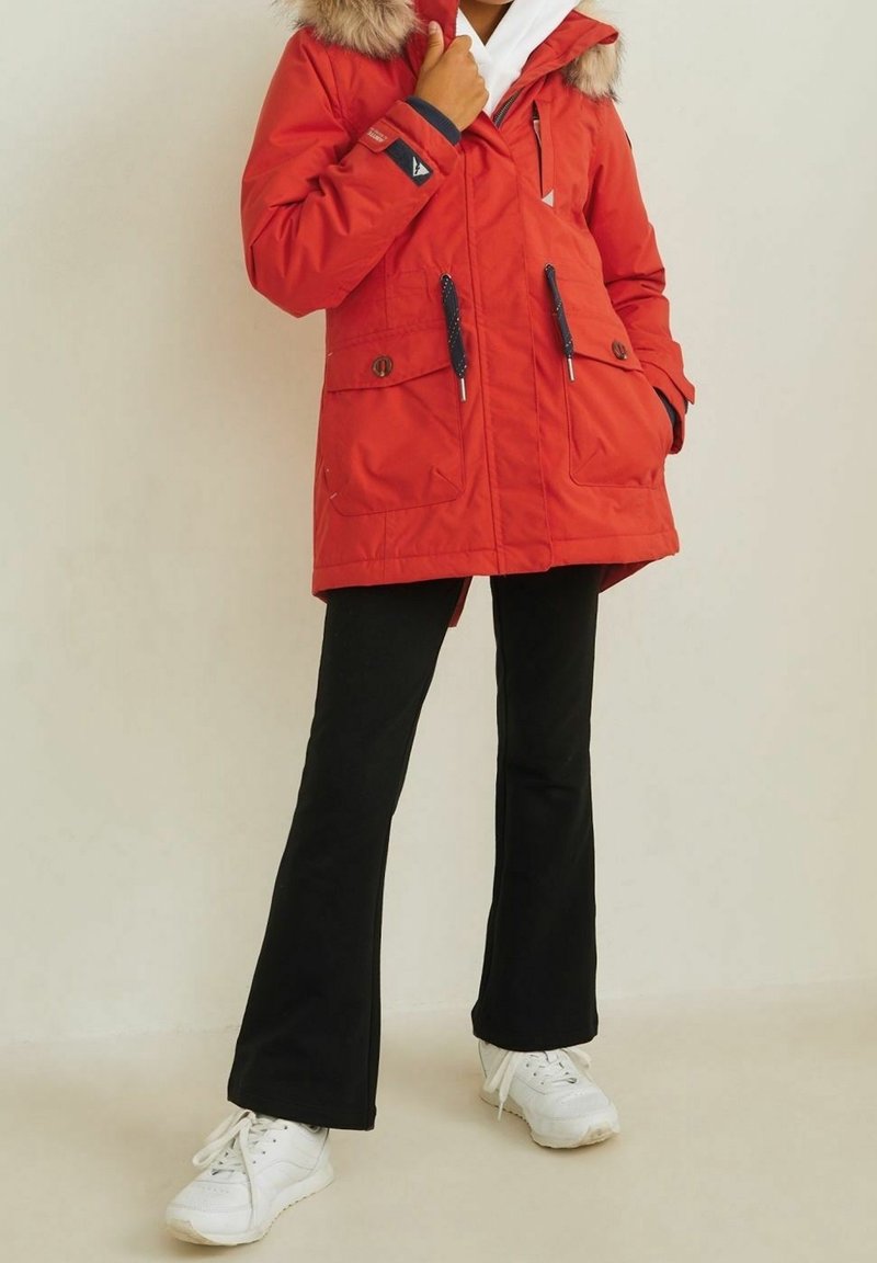 Czerwony ocieplany parka z kapturem sztucznym futrem, dwiema przednimi kieszeniami, elastycznymi mankietami i sznurkiem w talii. Połączona z czarnymi spodniami dzwonami i białymi trampkami.