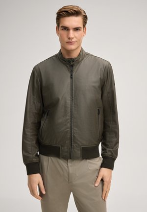 Giovane uomo in piedi, indossa una giacca bomber verde oliva con zip e pantaloni beige chiaro, su uno sfondo neutro uniforme.