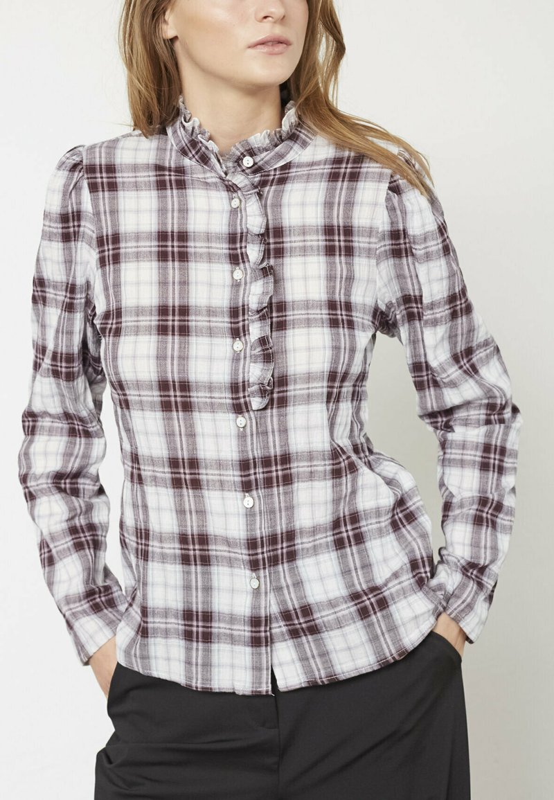 Kvinde iført en hvid og burgunderternet, rynket skjorte med knapper og sorte bukser, hænderne i lommerne, mod en ensfarvet lys baggrund.