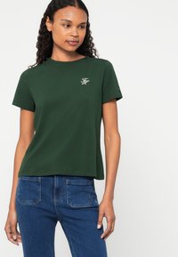 Πράσινο βαμβακερό t-shirt με στρογγυλή λαιμόκοψη και κοντά μανίκια. Διαθέτει διακριτό κεντητό λογότυπο στο στήθος. Συνδυάζεται με σκούρο μπλε τζιν παντελόνι.