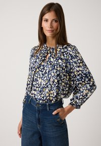 Blumenbluse in Marineblau mit gelben und blauen Blumen, mit gebundenem Ausschnitt und Puffärmeln, kombiniert mit hoch taillierten Jeans.