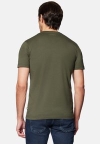 Oliwkowa koszulka z krótkim rękawem, okrągłym dekoltem, gładką teksturą i delikatnym logo marki z tyłu. Noszona z ciemnoniebieskimi dżinsami.