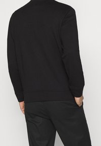 Felpa nera con polsini e orlo a costine, caratterizzata da un collo rotondo e una texture di tessuto liscia. Indossata con pantaloni neri.