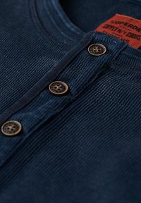 Navy blue ribbed knit shirt with three brown buttons and an orange label showing brand and product details inside collar.