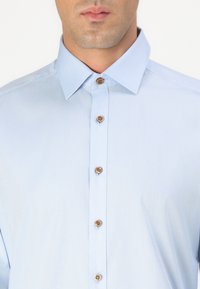 Lichtblauw overhemd met knopen van een gestructureerde stof, met een puntige kraag en vier bruine knoopsluitingen aan de voorkant.