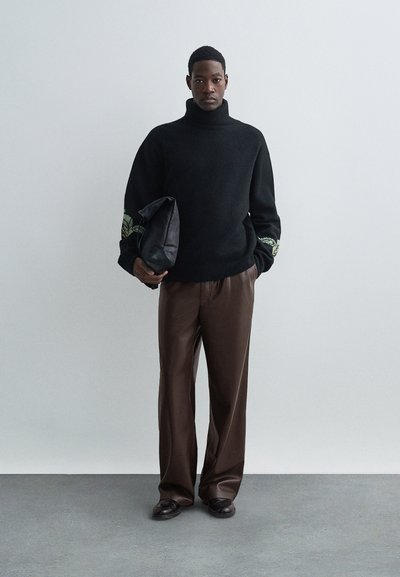 Tânăr îmbrăcat cu un pulover negru cu guler înalt și mâneci florale, pantaloni lejeri maro, pantofi întunecați, ținând o geantă pliabilă neagră.