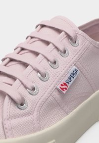 Ανοιχτό ροζ καμβά αθλητικό παπούτσι με κορδόνια στο ίδιο χρώμα, μεταλλικά ροντέλα, λευκή καουτσούκ σόλα και μια ταμπέλα με τη μάρκα Superga στο πλάι.