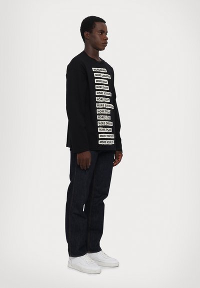 Black long-sleeve shirt with white text boxes featuring various phrases, paired with dark jeans and white sneakers. Simple design, relaxed fit.