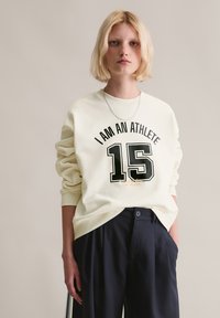 Kremowy sweatshirt z czarnym nadrukiem "JESTEM SPORTOWCEM 15", ze ściągaczami i okrągłym dekoltem. W zestawie z ciemnymi, plisowanymi spodniami. Srebrny naszyjnik.