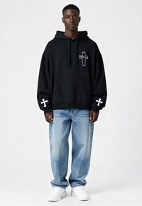 Felpa nera oversize con logo croce bianca sul petto e sulla manica. Abbinata a jeans baggy blu chiaro e sneakers bianche. Texture liscia.
