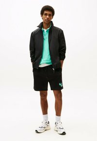 Zwarte ritsjack, turquoise poloshirt, zwarte shorts met groene grafische print, witte sokken en witte sneakers. Casual sportoutfit.
