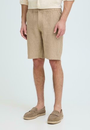 Modello maschile che indossa pantaloncini in lino beige con bottoni frontali e vita elastica, abbinati a scarpe beige abbinate senza lacci.