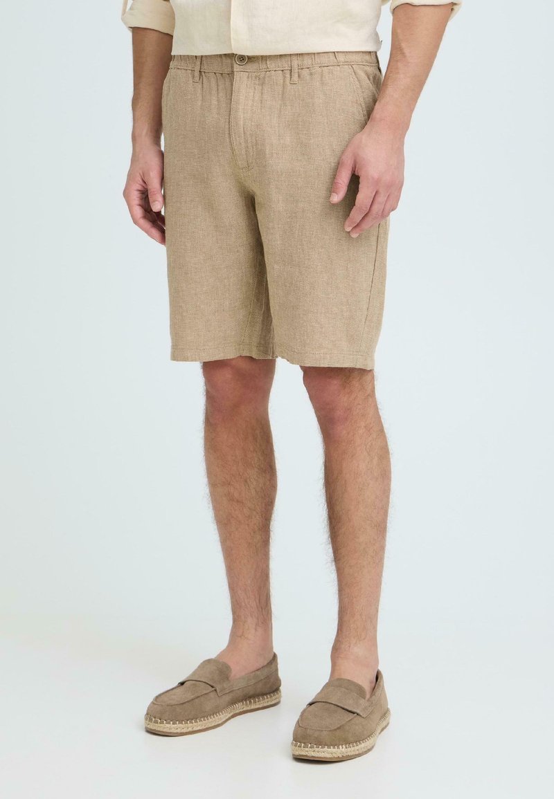Modèle masculin portant un short en lin beige avec bouton frontal et taille élastique, associé à des chaussures beige assorties à enfiler.