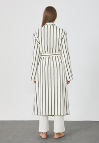 Csíkos köntös fehér alapszínen, fekete függőleges vonalakkal, texturált anyagból készült, hosszú ujjakkal és övvel. Fehér nadrág fölött viselve.