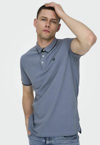 Polo de color gris claro con cuello, tres botones y ribete negro en contraste. Presenta un pequeño logo y mangas cortas.