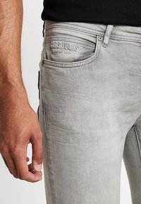 Primer plano de una persona que lleva puestos unos jeans gris claro con costuras visibles y un parche de la marca cerca del bolsillo delantero en la cadera derecha.