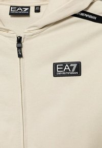 Felpa con cerniera color beige chiaro realizzata in tessuto morbido. Presenta un'etichetta con logo nera e un cappuccio con cordino. Include una cerniera centrale.