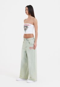 Witte strapless top met kleurrijke grafische prints; lichtgroene wijde jeans met rode geborduurde logo; witte sneakers.