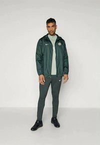 Green lightweight jacket with hood, white Nike logo, worn over a light green shirt. Slim-fit dark green pants and black athletic shoes.
