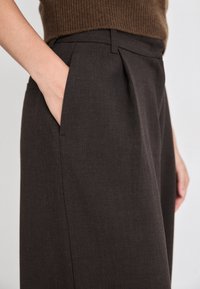 Pantaloni marroni plissettati con una texture liscia, dotati di tasche laterali e una vestibilità su misura, mostrati dalla vita alla coscia.