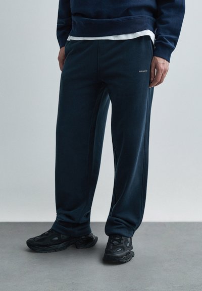 Pantaloni jogger blu navy con una texture morbida, caratterizzati da cuciture laterali e un piccolo logo sulla coscia. Abbinati a scarpe sportive nere.
