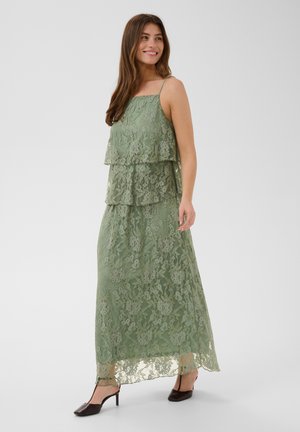 Uśmiechnięta kobieta w warstwowej zielonej maksi sukience z koronką, z cienkimi ramiączkami i czarnych sandałkach na obcasie typu slingback, stojąca na jednolitym tle.