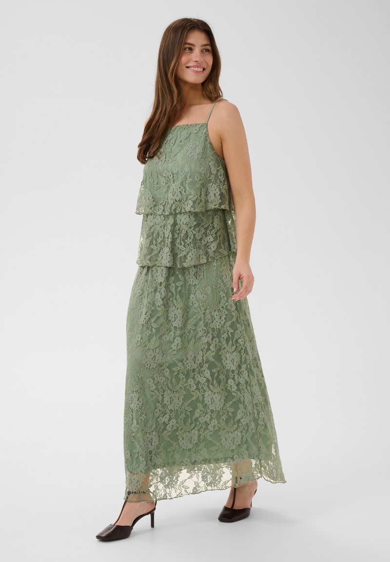Femme souriante portant une robe maxi en dentelle verte superposée avec de fines bretelles et des chaussures slingback noires à talons, debout devant un fond uni.