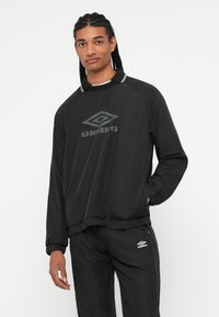 Noor mees, kellel on punutud juuksed, kannab musta Umbro treeningkostüümi, millel on logo prinditud nii särgile kui pükstele, seismas lihtsa tausta ees.
