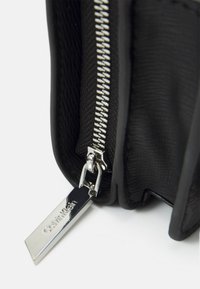 Zbliżenie na czarny zamek błyskawiczny torby o teksturowanej powierzchni z srebrnym uchwytem z logo Calvin Klein na białej powierzchni.