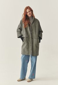 Cappotto di lana grigio con grandi tasche a toppa, chiusura a bottone e vestibilità comoda. Abbinato a jeans azzurri ricamati e scarpe beige.