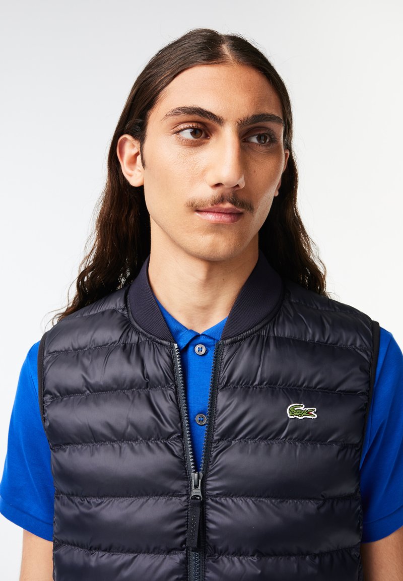 Doudoune navy sans manches avec fermeture éclair à l'avant, col côtelé et logo Lacoste. Portée par-dessus un polo bleu, à manches courtes et avec deux boutons.
