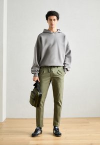 Hanorac gri cu un croi lejer, pantaloni de culoare verde măslin, pantofi atletici negri și o geantă de umăr neagră, așezate pe un fundal neutru.