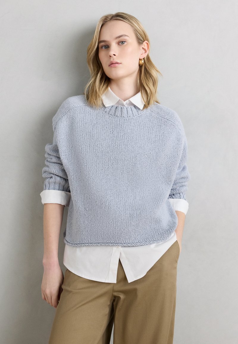 Νεαρή γυναίκα με ξανθά μαλλιά, φορώντας χρυσούς κρίκους στα σκουλαρίκια, ανοιχτό μπλε πλεκτό πουλόβερ πάνω από λευκό πουκάμισο και μπεζ παντελόνι, με γκρι φόντο.