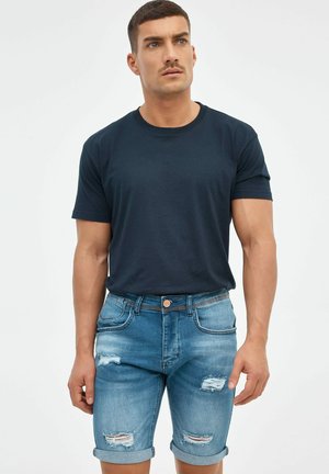 Camiseta de algodón azul marino con cuello redondo, combinada con unos pantalones cortos de mezclilla azul claro desgastados con dobladillos enrollados y costuras decorativas.