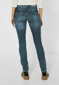 Jeans de mezclilla azul oscuro con un ajuste slim, que cuentan con dos bolsillos traseros y un ligero desgaste. Combinados con zapatos negros y una parte superior en tonos neutros.