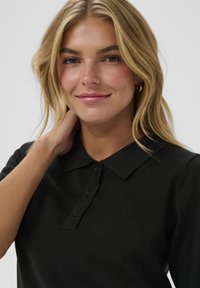 Polo de color negro con cuello, que presenta una tapeta de botones. La tela tiene una textura suave, complementando el diseño sencillo.