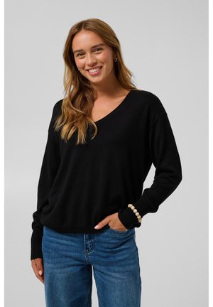 Jeune femme souriante portant un haut noir à manches longues, un jean bleu et un bracelet blanc en perles, debout devant un fond neutre.