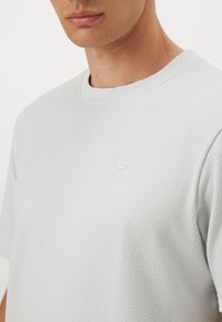 Lys grå Nike T-shirt med et struktureret, vaffelmønstret stof, rund halsudskæring og et lille logo på brystet. Korte ærmer.