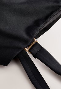 Gros plan d’un tissu noir avec bords cousus et sangle réglable avec une boucle métallique rectangulaire dorée sur un fond uni.