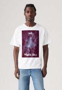 Mand iført en hvid T-shirt med et tryk af en lilla skov og stjernehimmel samt blå jeans, stående foran en ensfarvet lys baggrund.