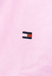 Kleines rechteckiges gesticktes Logo in Rot, Weiß und Schwarz auf hellrosa strukturiertem Stoffhintergrund.