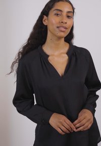 Blusa nera a maniche lunghe con scollatura a V profonda, texture morbida, dettagli plissettati al collo e polsini con bottoni. Tessuto liscio e fluente.