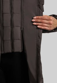Mano che svuota una tasca della giacca imbottita marrone scuro, mostrando numerosi zip verticali e dettagli di cucitura trapuntati.