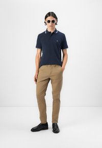 Jūras spēku polo krekls ar baltu apkakles apdari, khaki bikses un melnas kurpes. Modelis valkā saulesbrilles, ieņemot neitrālu pozīciju uz baltas fona.