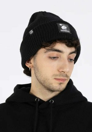 Nuori mies, jolla on vaalea parta, pukeutuneena mustaan neulottuun pipoon ja mustaan huppariin, katsoo alas ja sivulle yksinkertaista taustaa vasten.